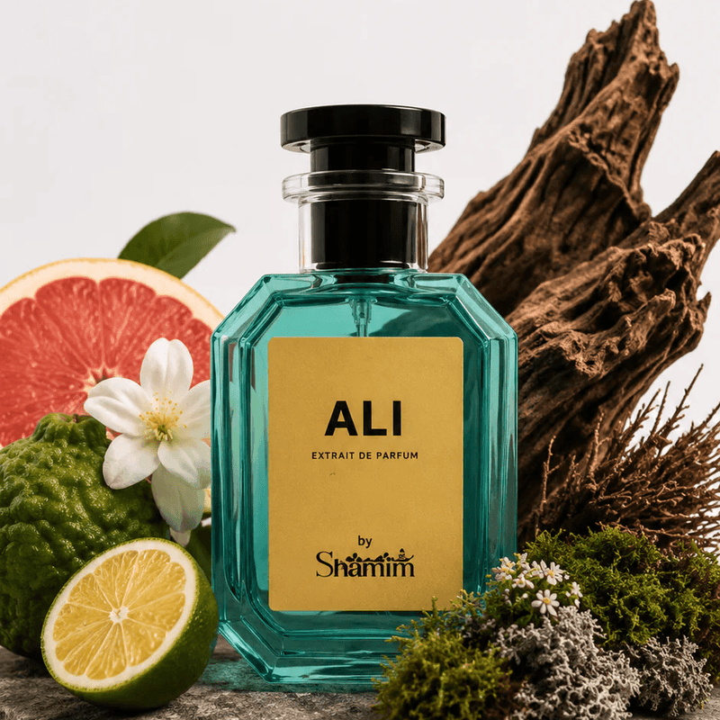 A faceted teal glass perfume bottle with a black cap and a gold label reading 'ALI' and 'by Saintri' is styled on a stone slab with citrus slices, a white flower, moss, and driftwood against a clean white background.