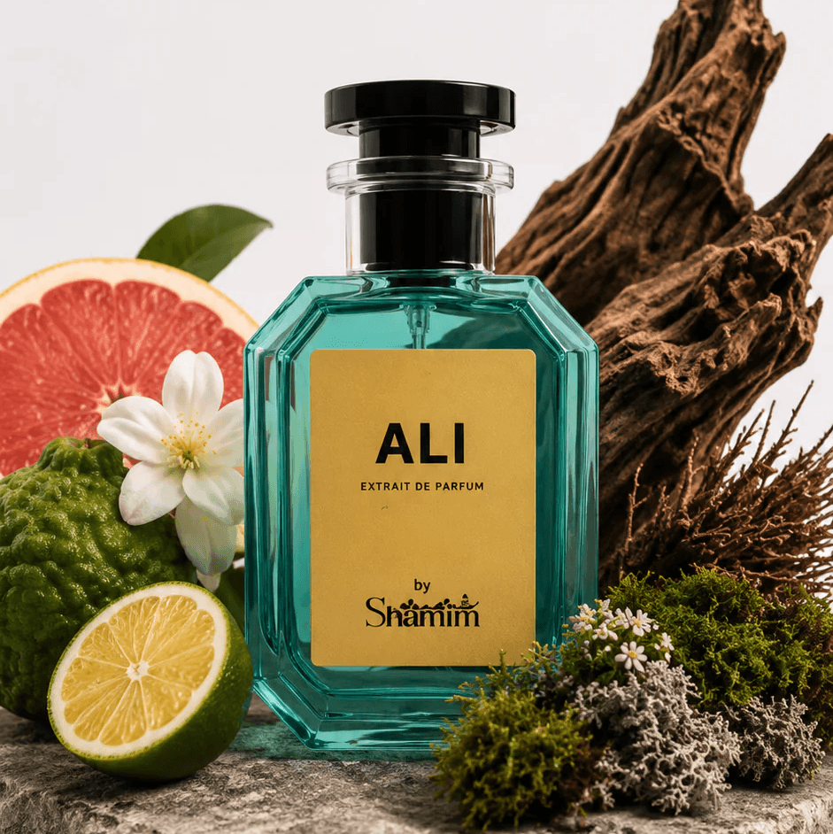 A faceted teal glass perfume bottle with a black cap and a gold label reading 'ALI' and 'by Saintri' is styled on a stone slab with citrus slices, a white flower, moss, and driftwood against a clean white background.
