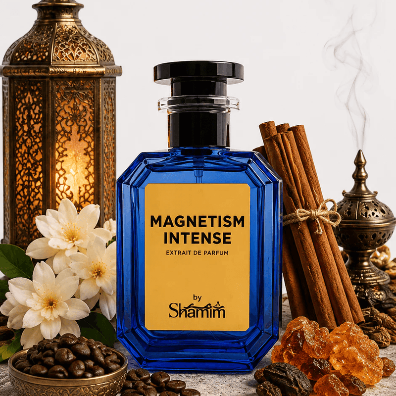 A blue glass perfume bottle labeled 'MAGNETISM INTENSE' by Shamin, styled on a stone slab with cinnamon sticks, coffee beans, white flowers, amber crystals, and brass lanterns against a light gray background.