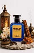 A blue glass perfume bottle labeled 'MAGNETISM INTENSE' by Shamin, styled on a stone slab with cinnamon sticks, coffee beans, white flowers, amber crystals, and brass lanterns against a light gray background.
