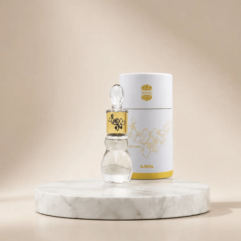 A small clear glass dropper bottle with a gold label stands in front of a white cylindrical product box with gold detailing, both placed on a round white marble pedestal against a warm beige studio background.