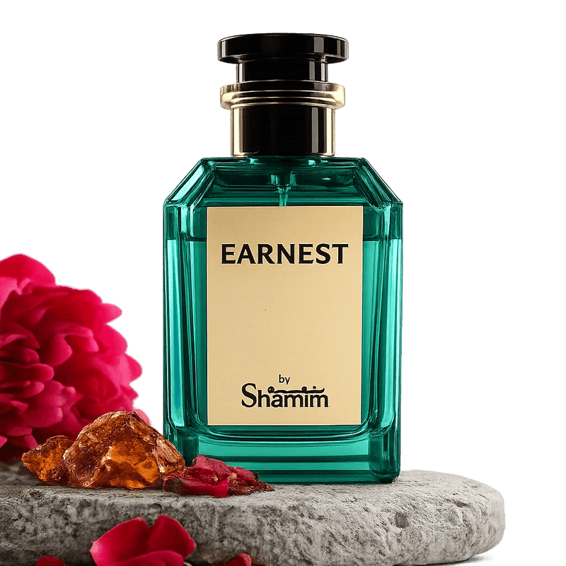 A faceted teal-green perfume bottle labeled 'EARNEST by Shamim' with a cream label and black cap, displayed on a stone slab with red flowers and amber stones against a black background.