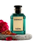 A faceted teal-green perfume bottle labeled 'EARNEST by Shamim' with a cream label and black cap, displayed on a stone slab with red flowers and amber stones against a black background.