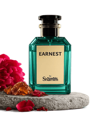 A faceted teal-green perfume bottle labeled 'EARNEST by Shamim' with a cream label and black cap, displayed on a stone slab with red flowers and amber stones against a black background.