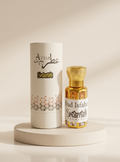A bottle of Attar Skamim oud perfume next to its white cylindrical packaging box, displayed on a round beige pedestal against a warm beige background.