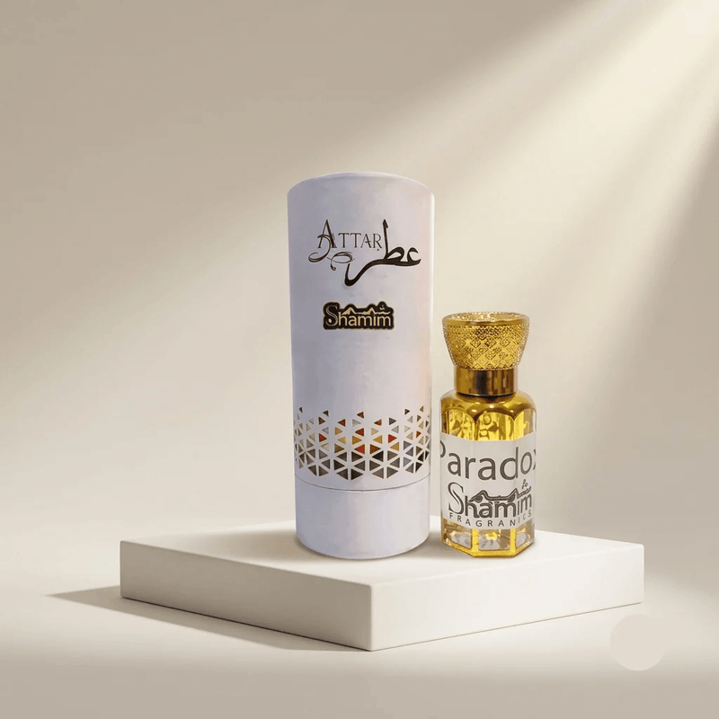 A small bottle of Attar Shammim perfume with a gold cap stands next to its tall white cylindrical box on a white pedestal. The bottle contains a golden-yellow liquid, and the box is white with black and gold branding.