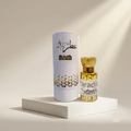 A small bottle of Attar Shammim perfume with a gold cap stands next to its tall white cylindrical box on a white pedestal. The bottle contains a golden-yellow liquid, and the box is white with black and gold branding.
