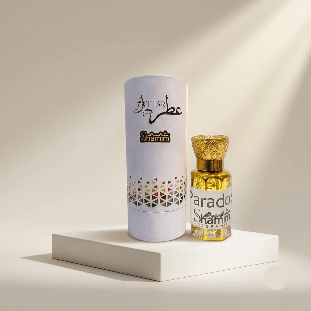 A small bottle of Attar Shammim perfume with a gold cap stands next to its tall white cylindrical box on a white pedestal. The bottle contains a golden-yellow liquid, and the box is white with black and gold branding.