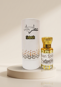 A small ornate bottle with a gold cap and gold-toned liquid beside a tall white cylindrical box, both printed with the brand name Attar Scamim, displayed on a round beige pedestal against a warm cream background.