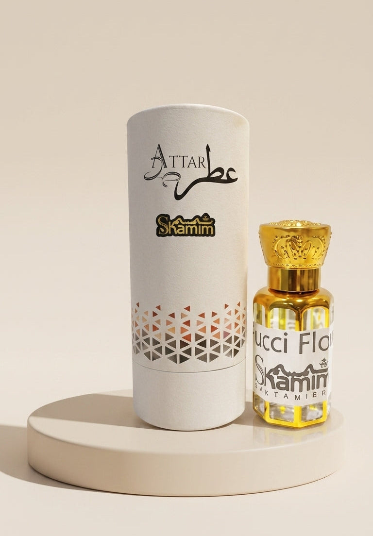 Attar Skamin perfume bottle with golden liquid and cap next to its white cylindrical packaging box, displayed on a round beige pedestal against a warm beige background.