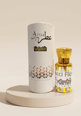 Attar Skamin perfume bottle with golden liquid and cap next to its white cylindrical packaging box, displayed on a round beige pedestal against a warm beige background.