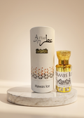 A white cylindrical box labeled 'ATTAR Oman' and 'Hawas Ice' stands beside a small faceted gold perfume bottle with a gold cap on a round white marble pedestal against a warm beige background.