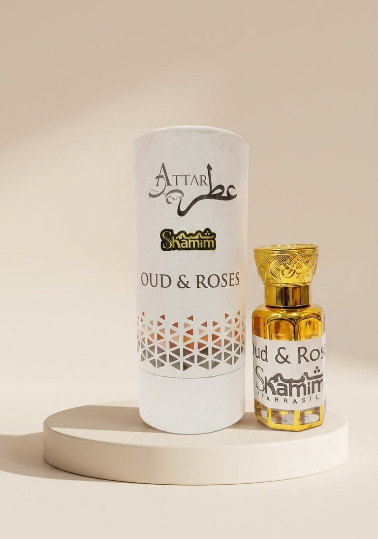 A white cylindrical perfume packaging tube and a small gold-toned bottle labeled 'Attar Shammim Oud & Roses' displayed on a round pedestal against a warm beige background.