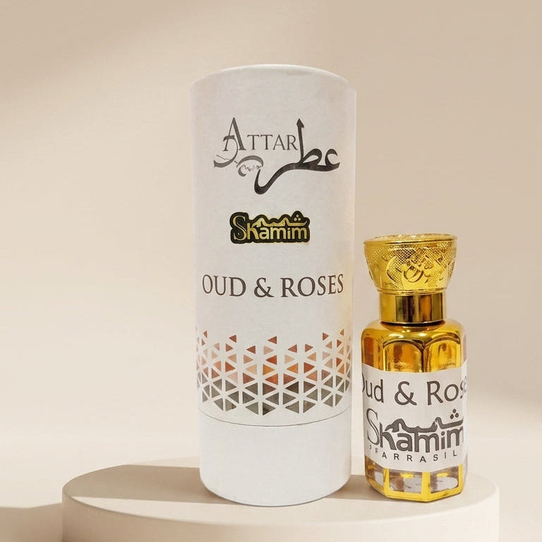 A white cylindrical perfume packaging tube and a small gold-toned bottle labeled 'Attar Shammim Oud & Roses' displayed on a round pedestal against a warm beige background.