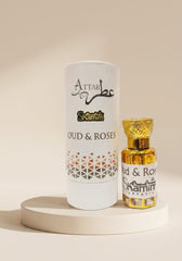 A white cylindrical perfume packaging tube and a small gold-toned bottle labeled 'Attar Shammim Oud & Roses' displayed on a round pedestal against a warm beige background.