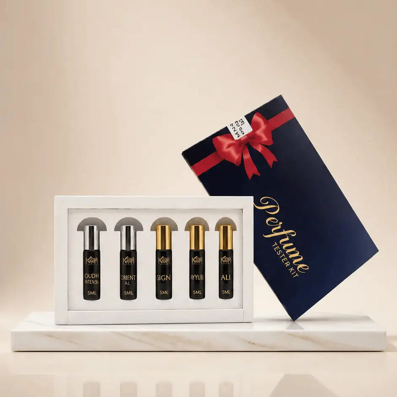 A perfume gift set featuring four small black spray bottles with gold caps in a white tray, alongside a dark navy blue gift box with a red ribbon and gold 'Perfume' lettering, set on a marble-like base against a warm beige background.
