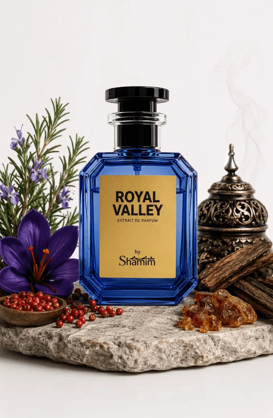 A faceted cobalt blue perfume bottle with a black cap and a gold label reading 'ROYAL VALLEY' and 'by Shammim', styled on a stone slab with a purple flower, red berries, greenery, amber pieces, and a metal incense burner against a white background.