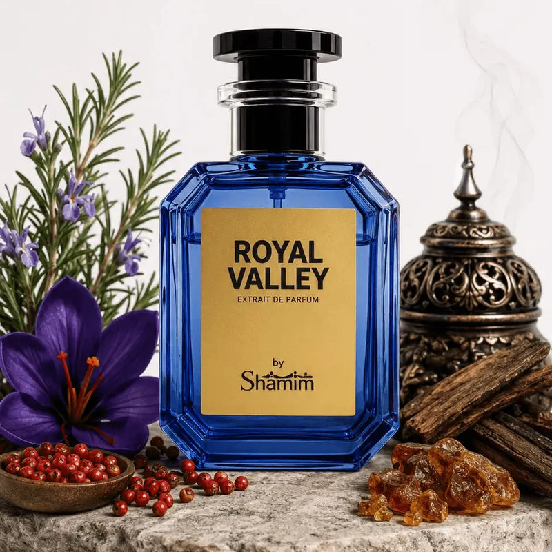 A faceted cobalt blue perfume bottle with a black cap and a gold label reading 'ROYAL VALLEY' and 'by Shammim', styled on a stone slab with a purple flower, red berries, greenery, amber pieces, and a metal incense burner against a white background.