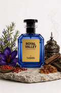 A faceted cobalt blue perfume bottle with a black cap and a gold label reading 'ROYAL VALLEY' and 'by Shammim', styled on a stone slab with a purple flower, red berries, greenery, amber pieces, and a metal incense burner against a white background.