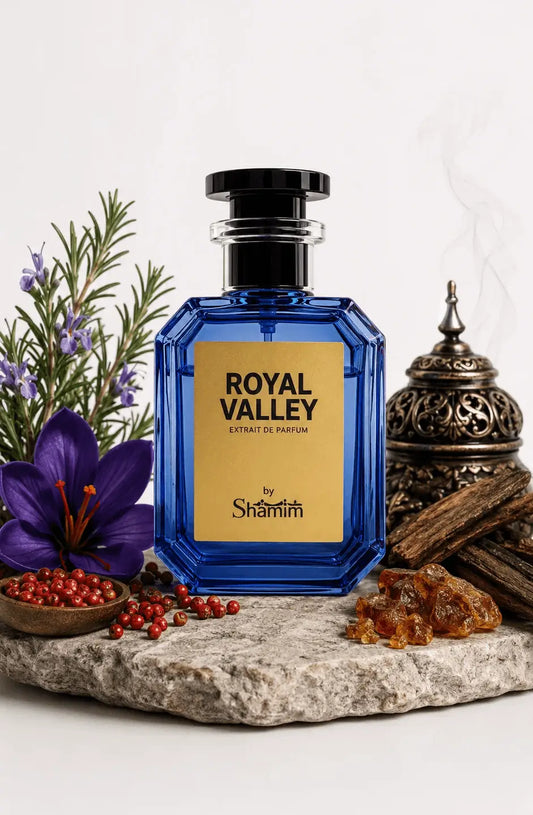 A faceted cobalt blue perfume bottle with a black cap and a gold label reading 'ROYAL VALLEY' and 'by Shammim', styled on a stone slab with a purple flower, red berries, greenery, amber pieces, and a metal incense burner against a white background.
