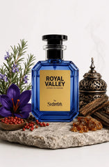A faceted cobalt blue perfume bottle with a black cap and a gold label reading 'ROYAL VALLEY' and 'by Shammim', styled on a stone slab with a purple flower, red berries, greenery, amber pieces, and a metal incense burner against a white background.