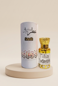 A small glass bottle of Attar Shammim fragrance oil with a gold cap stands next to its tall white cylindrical packaging tube on a round beige pedestal. The tube is printed with the brand name 'Attar Shammim' and a geometric pattern near the bottom.