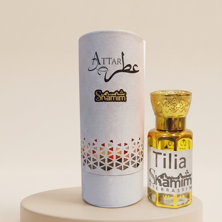 A small glass bottle of Attar Shammim fragrance oil with a gold cap stands next to its tall white cylindrical packaging tube on a round beige pedestal. The tube is printed with the brand name 'Attar Shammim' and a geometric pattern near the bottom.