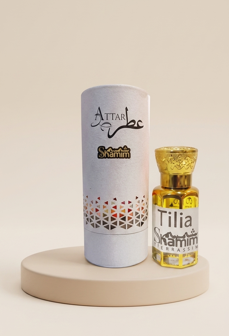 A small glass bottle of Attar Shammim fragrance oil with a gold cap stands next to its tall white cylindrical packaging tube on a round beige pedestal. The tube is printed with the brand name 'Attar Shammim' and a geometric pattern near the bottom.