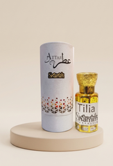 A small glass bottle of Attar Shammim fragrance oil with a gold cap stands next to its tall white cylindrical packaging tube on a round beige pedestal. The tube is printed with the brand name 'Attar Shammim' and a geometric pattern near the bottom.