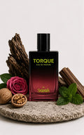 A bottle of Torque Eau de Parfum by Skaramell, featuring a black-to-deep-red gradient glass bottle with gold lettering, styled on a stone plate with a rose, walnuts, cinnamon, and mint leaves against a light gray background.