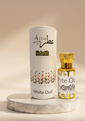 A cylindrical white perfume box labeled 'Attar Shammim White Oud' stands beside a small clear glass perfume bottle with a gold cap on a marble pedestal against a warm beige background.
