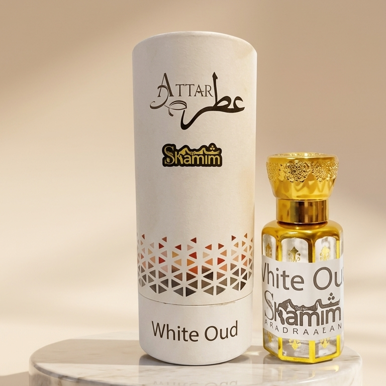 A cylindrical white perfume box labeled 'Attar Shammim White Oud' stands beside a small clear glass perfume bottle with a gold cap on a marble pedestal against a warm beige background.